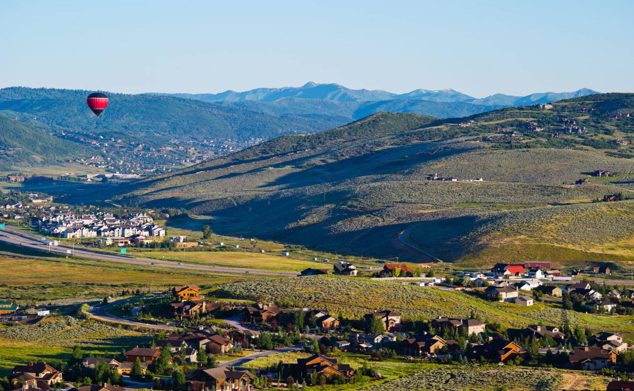 A red hot air balloon floats above a valley with scattered houses, green hills, and distant mountains under a clear blue sky.