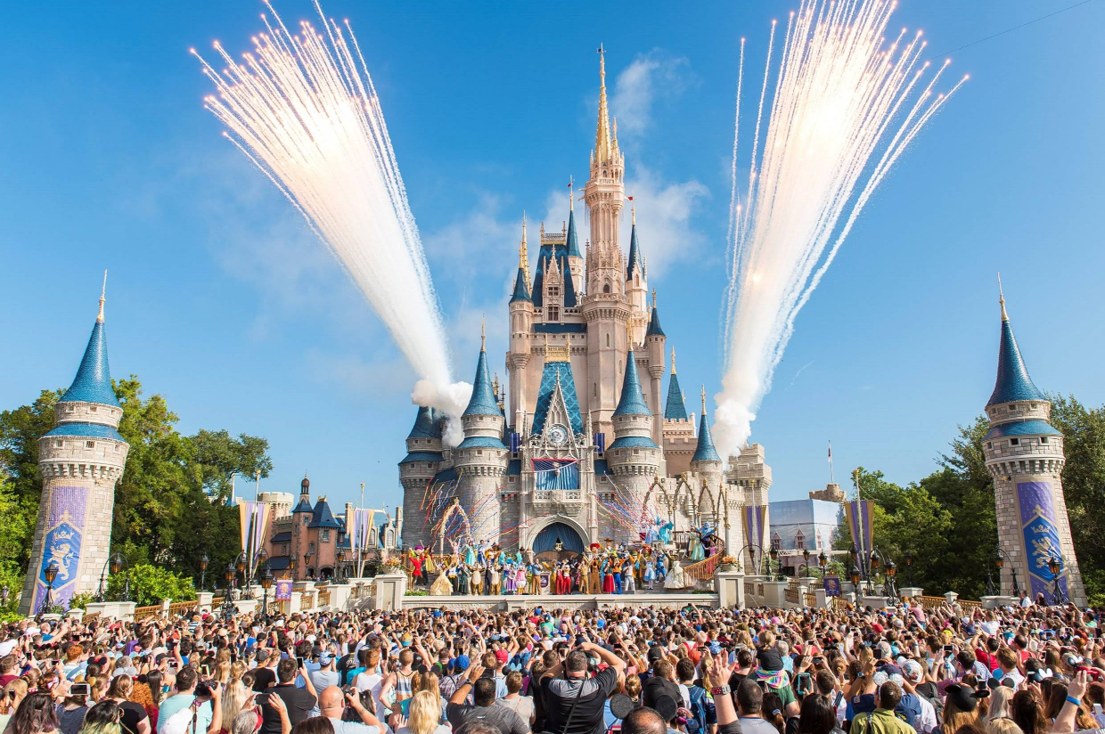 A large crowd gathers in front of Cinderella's Castle at Disney World as fireworks shoot into the blue sky and costumed characters perform on stage.