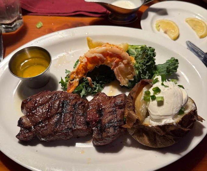 A dinner plate from a steakhouse on the West Coast with grilled steak, a lobster tail on greens, a baked potato topped with sour cream and chives, a cup of melted butter, and lemon wedges on the side.