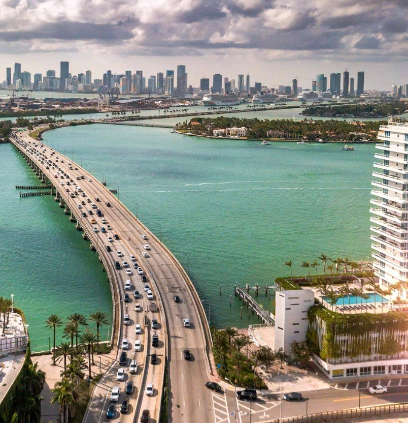 A wide bridge with multiple lanes of traffic crosses turquoise water toward a city skyline with tall buildings under a cloudy sky; palm trees and a swimming pool are in the foreground.