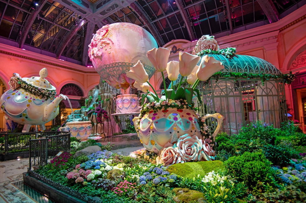 A whimsical indoor garden display, one of the charming Las Vegas attractions, features giant teapots, teacups with oversized white calla lilies, colorful flowers, and greenery under a glass ceiling with warm, pink-hued lighting.