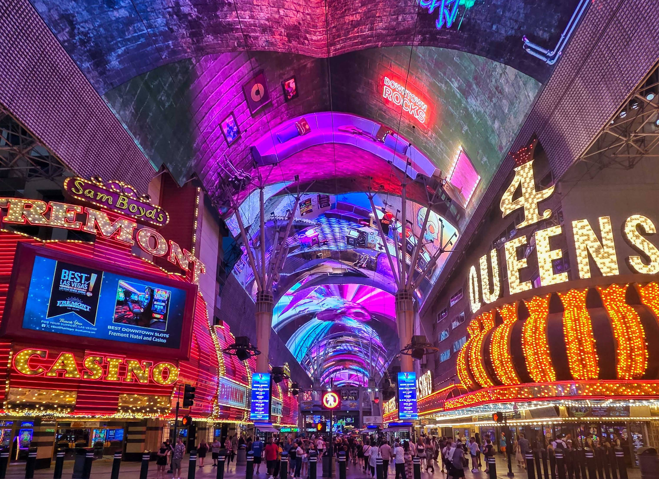 A vibrant street scene shows one of the top Las Vegas attractions—Fremont Street at night—filled with colorful neon casino signs like "4 Queens" and "Fremont," as crowds stroll beneath the dazzling, digital canopy.