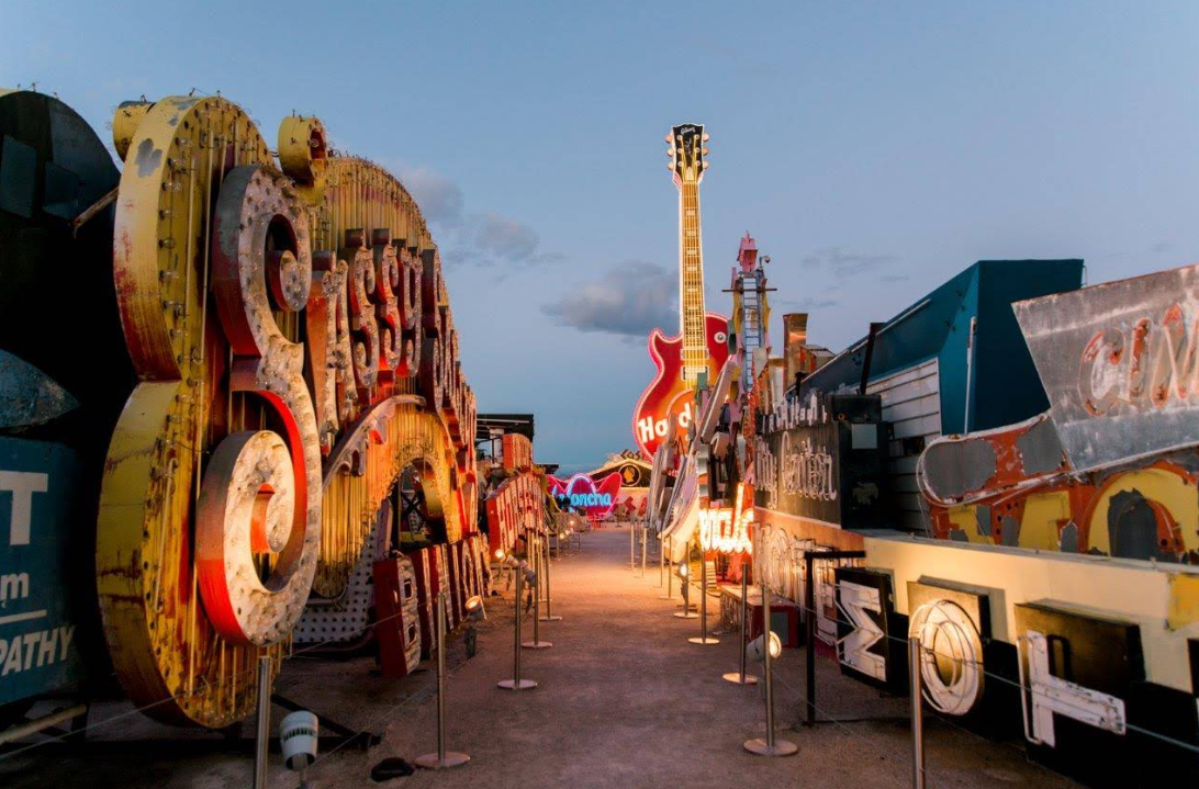 A walkway lined with large, colorful vintage neon signs—including guitars and old casino names—leads into the distance under a dusky sky at an outdoor museum, showcasing one of the must-see Las Vegas attractions.