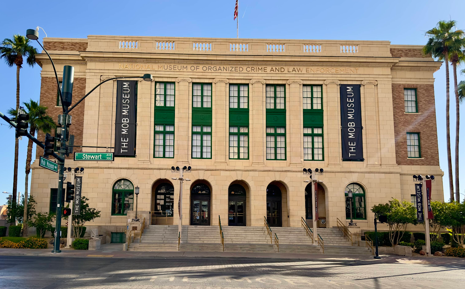 The Mob Museum, one of the top Las Vegas attractions, is a large beige stone building with tall arched windows, banners, and a grand staircase, standing on a street corner under a clear blue sky surrounded by palm trees.
