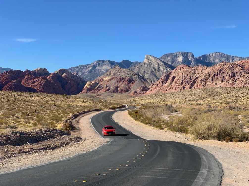 A red car drives along a winding road through a desert landscape with rocky hills and distant mountains, capturing the spirit of adventurous Las Vegas attractions under a clear blue sky.