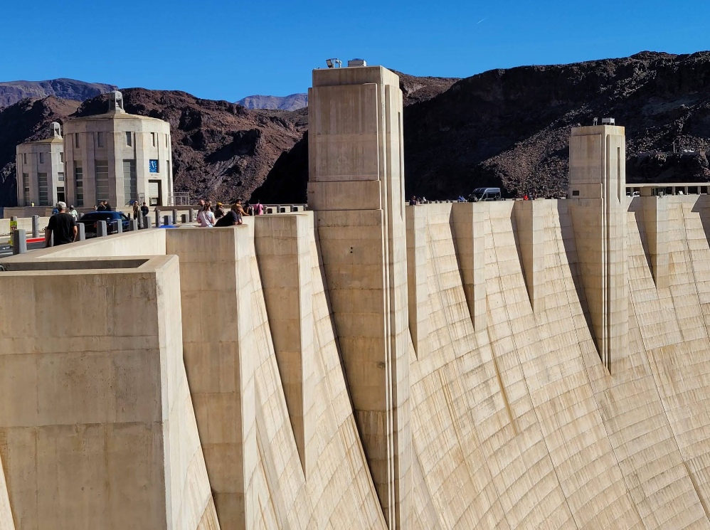 A large concrete dam, one of the iconic Las Vegas attractions, curves across a canyon as people stroll along the top. Rocky hills and blue sky frame the impressive structure in the background.