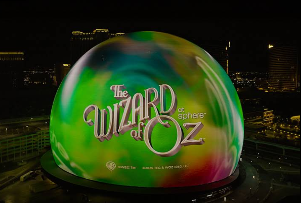 A large spherical building, one of the standout Las Vegas attractions, is lit up at night with swirling green, yellow, and pink colors and displays "The Wizard of Oz at Sphere" on its surface. City lights and buildings glow in the background.