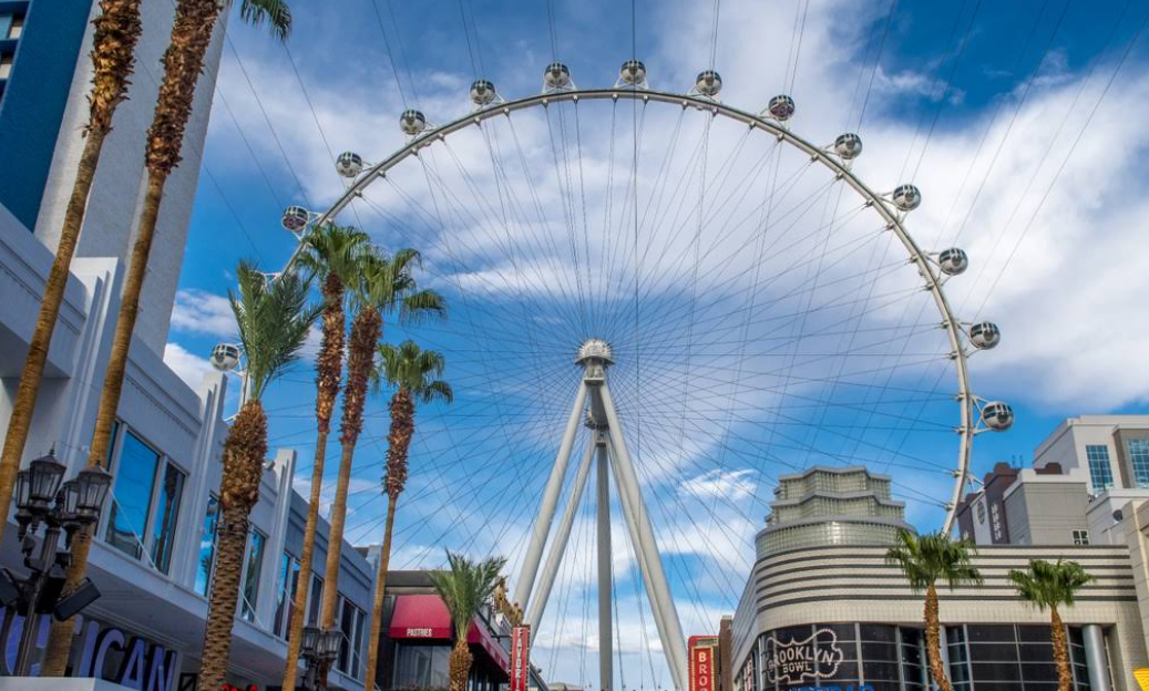 A large Ferris wheel, the High Roller—one of the top Las Vegas attractions—towers above palm trees and buildings under a blue sky with wispy clouds. Urban shops and neon lights line the bustling street below.