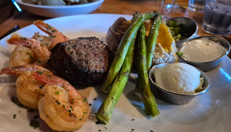 A plate at a steakhouse on the West Coast featuring grilled shrimp, a steak filet, roasted asparagus, and a baked potato topped with butter and cheese, plus sides of sour cream and white dipping sauce, served on a restaurant table.