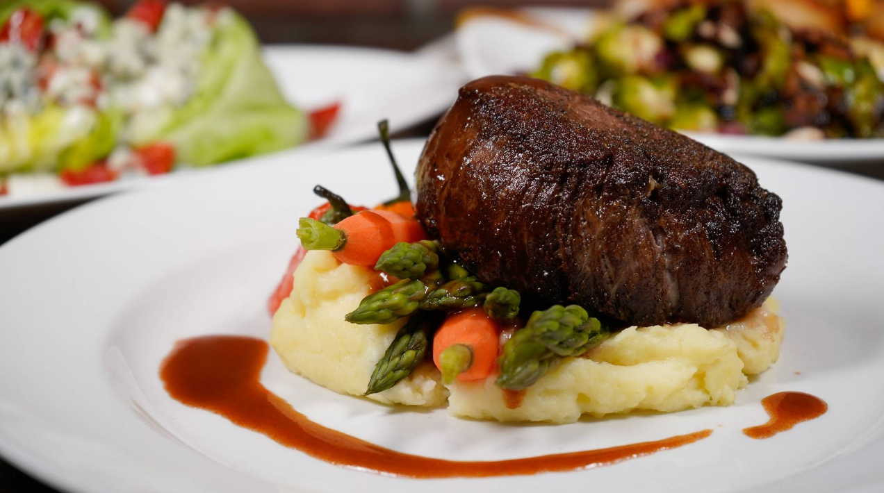 A seared filet mignon atop mashed potatoes, garnished with asparagus and carrots, drizzled with brown sauce—an elegant dish reminiscent of fine dining at top Las Vegas attractions, served on a white plate with blurred side dishes in the background.