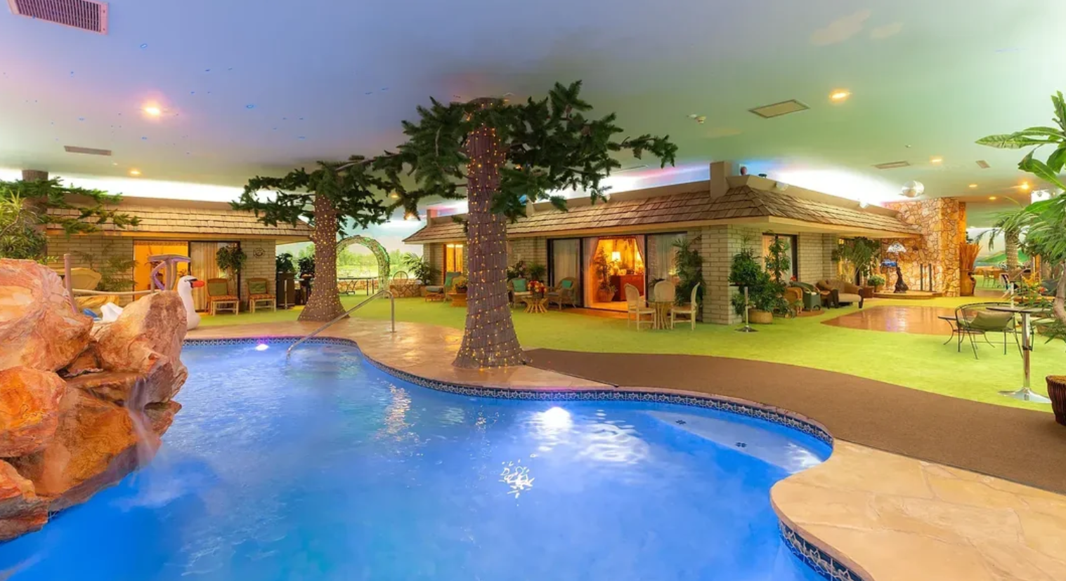 An indoor pool with palm trees, artificial grass, and a rock waterfall sits beside bungalow-style rooms under a painted sky ceiling, offering a tropical resort vibe reminiscent of top Las Vegas attractions.