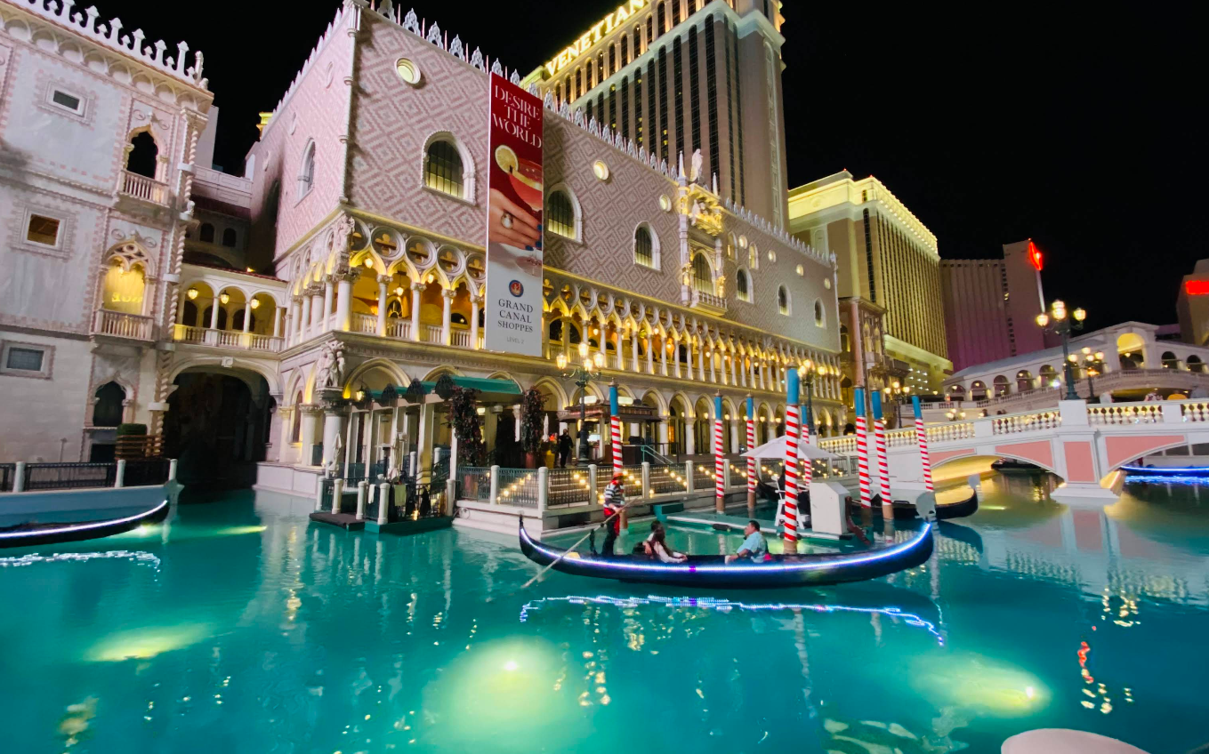A gondola floats on a turquoise canal in front of the Venetian hotel, one of the top Las Vegas attractions, featuring ornate, Italian-inspired architecture and striped poles, with glowing lights illuminating the night scene.