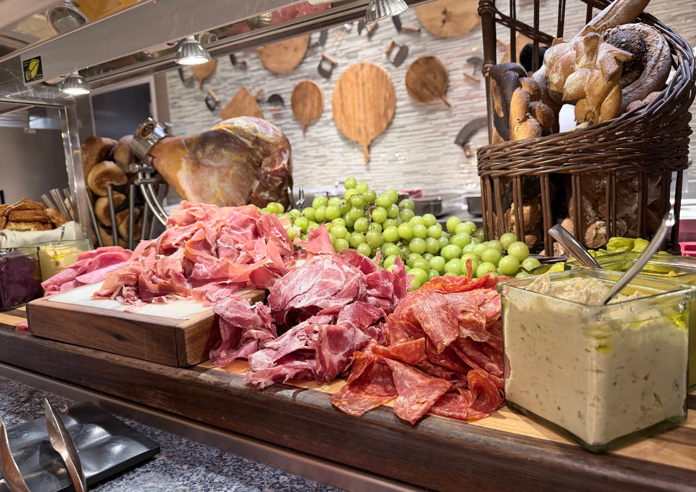 A buffet spread featuring assorted sliced deli meats, green grapes, a basket of breadsticks, and a dish of creamy dip; reminiscent of the vibrant feasts found at top Las Vegas attractions, with wooden utensils and a large ham decorating the background.