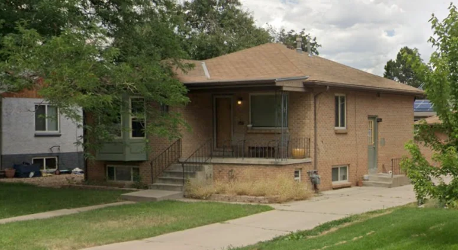 A single-story brick house from the early 1960s with a tan roof, small front porch with steps and railing, and a driveway. The green lawn has some dry patches, and mature trees surround this charming home built for living on a single income.