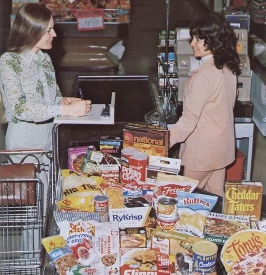 Two women stand at a grocery store checkout counter in the early 1960s on a single income, one as the cashier and the other as the customer. The cart is filled with various boxed and bagged food products, including chips, crackers, and cereals.