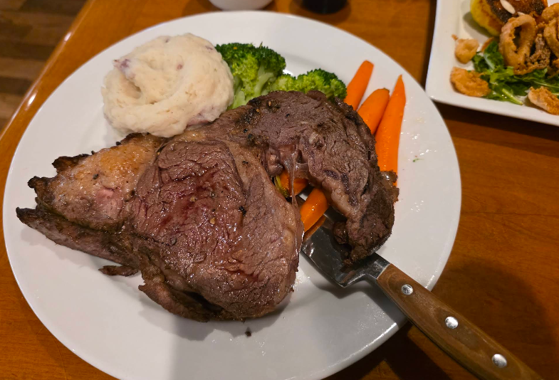 A plate with a large slice of roast beef, mashed potatoes, steamed broccoli, and carrots—classic fare at a steakhouse on the West Coast. A steak knife rests under the beef, while another dish with fried food appears in the background.