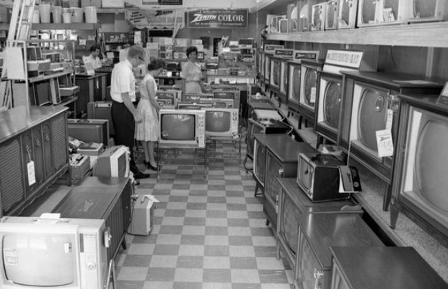 Black-and-white photo of a vintage electronics store in the early 1960s on a single income, with rows of old televisions. A man and woman stand together looking at TVs, while another person browses further back among shelves and displays.