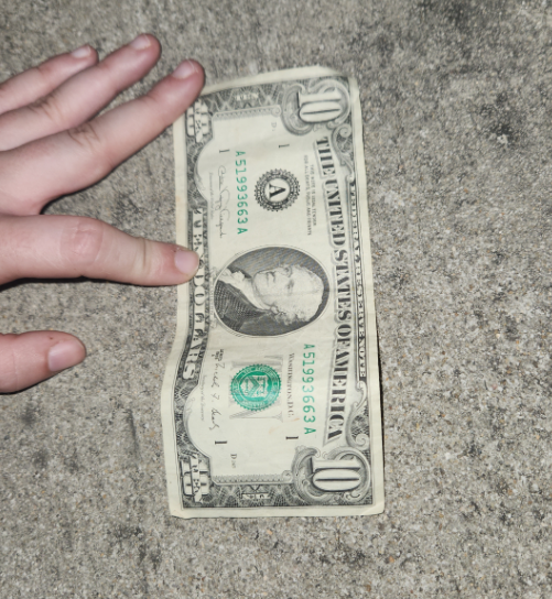 A person's left hand holds down a crumpled U.S. one-dollar bill on a rough, gray concrete surface at night, echoing the careful budgeting of the early 1960s on a single income.