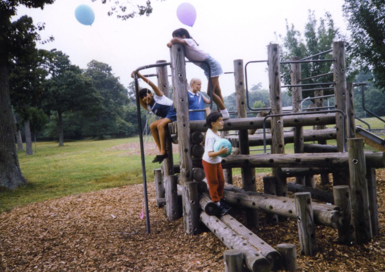 Four children play on a wooden playground structure in a park, echoing the carefree days of the early 1960s on a single income. Three climb and hang from the logs while one stands below holding a ball. Trees and balloons decorate the background.