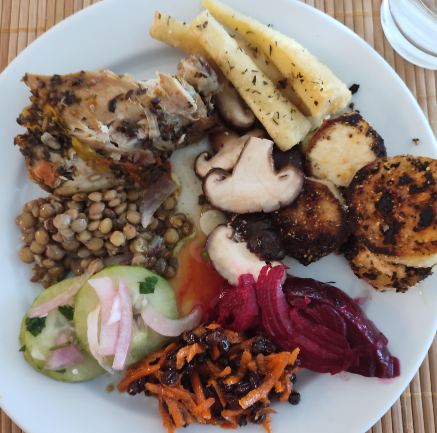 A white plate with roast chicken, roasted parsnips, sautéed mushrooms, breaded and fried rounds, lentil salad, cucumber salad, shredded beet, and a carrot-raisin salad—delicious ways to stretch food across several tasty sections.
