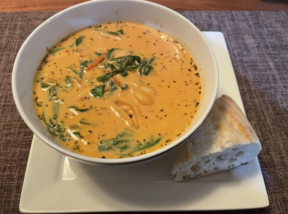 A bowl of creamy soup with pasta shells, greens, and herbs sits on a white plate alongside a piece of crusty bread on a brown placemat—perfect for stretching food into a hearty, satisfying meal.