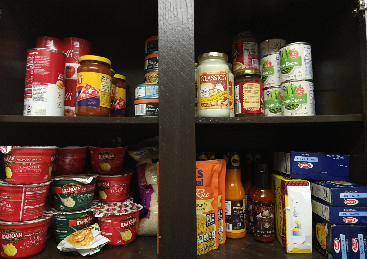 A pantry with shelves holding canned vegetables, pasta sauce jars, soup cans, instant mashed potatoes, hot sauces, dry pasta boxes, rice, and snacks—all neatly organized in a dark wooden cabinet to help stretch food for longer-lasting meals.