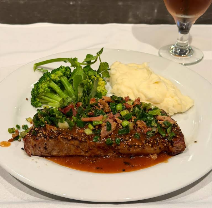 A plate of steakhouse on the west coast-style steak topped with chopped green onions and herbs in sauce, served with steamed broccoli and mashed potatoes. A glass of pink beverage is in the background.