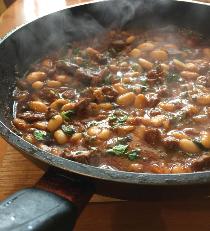 A steaming pan filled with a hearty stew of beef chunks, white beans, and chopped herbs in a rich, brown sauce sits on a wooden table—an ideal way to stretch food for nourishing family meals.