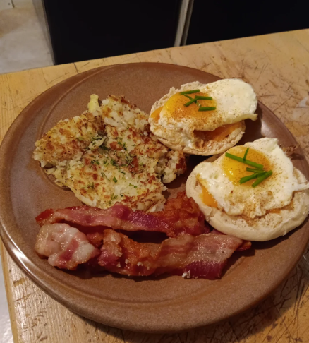A brown plate with crispy bacon strips, a golden hash brown patty topped with herbs, and two English muffins topped with fried eggs and chives offers a delicious way to stretch food on a cozy wooden table.