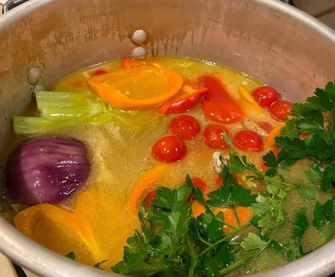 A pot of soup simmering with visible ingredients—cherry tomatoes, orange bell pepper slices, celery, a red onion chunk, and fresh parsley—simmers in a clear broth, the perfect way to stretch food into a hearty, flavorful meal.