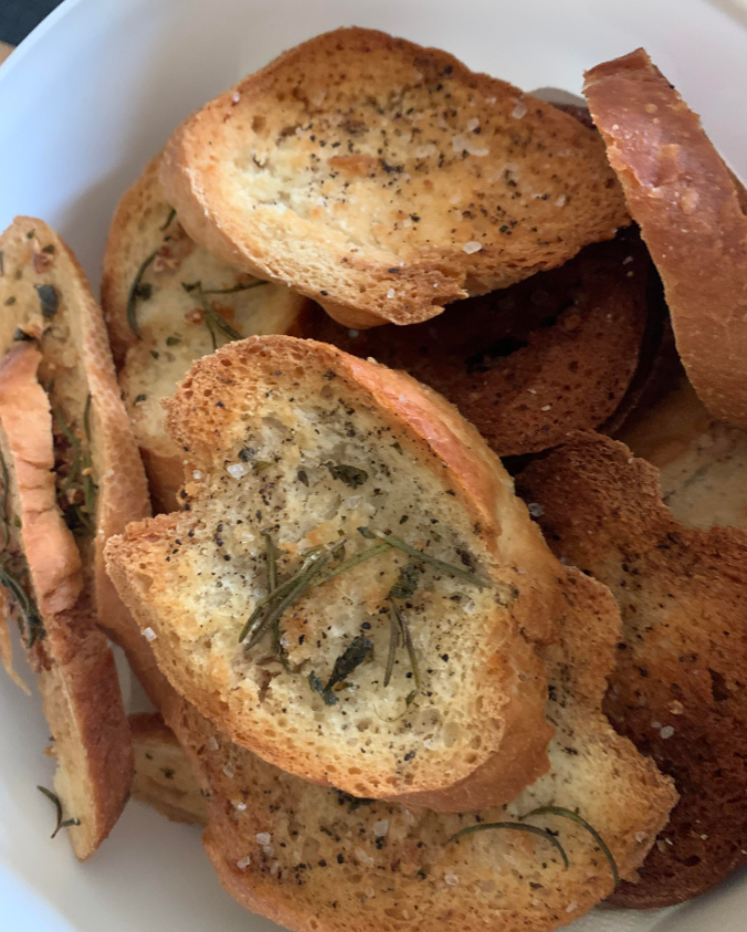 A close-up of several slices of toasted bread, perfect to stretch food, seasoned with black pepper, coarse salt, and fresh rosemary, arranged in a white bowl.