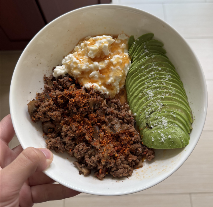 A hand holds a bowl with ground beef topped with seasoning, sliced avocado sprinkled with salt, and a scoop of cottage cheese with hot sauce—a delicious way to stretch food. The bowl is seen from above against a light background.