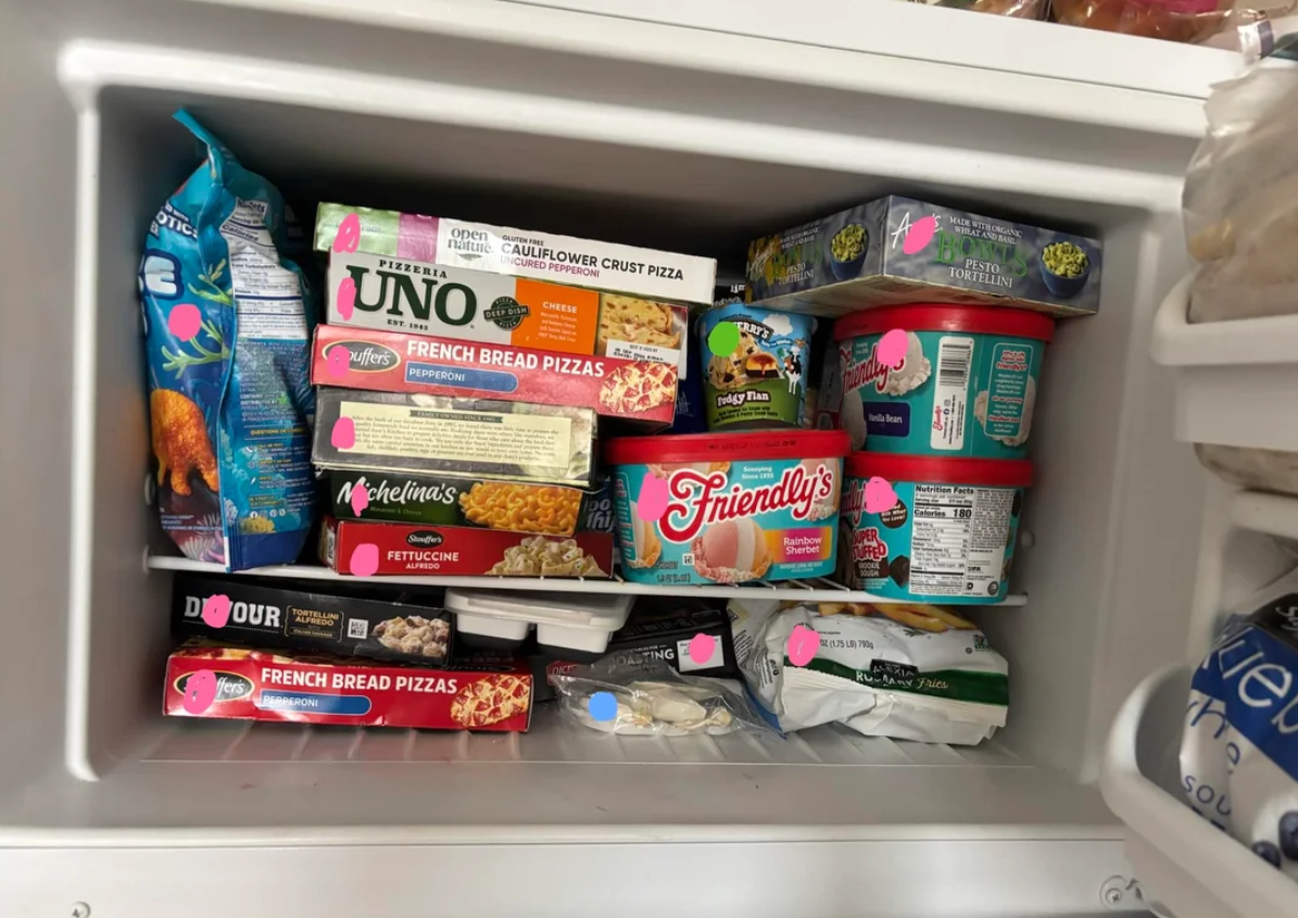 A packed freezer filled with various frozen foods, including pizzas, ice cream, pasta, and vegetables—many labeled with bright pink sticker dots to help stretch food supplies and keep meals organized.