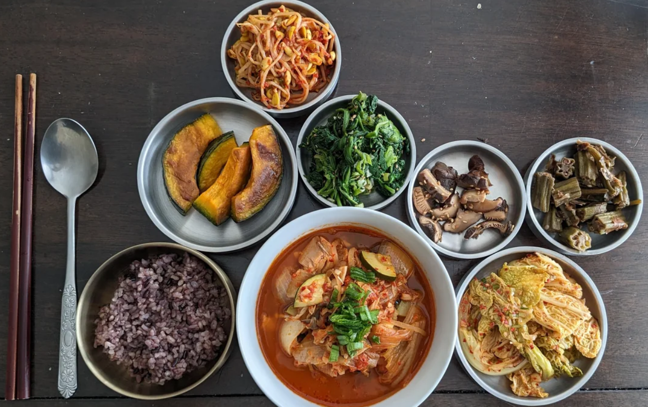 A Korean meal is arranged on a wooden table, featuring bowls of purple rice, kimchi stew, sautéed spinach, pumpkin, mushrooms, okra, bean sprout salad, and napa cabbage kimchi—perfect dishes to stretch food for a nourishing and satisfying dining experience.