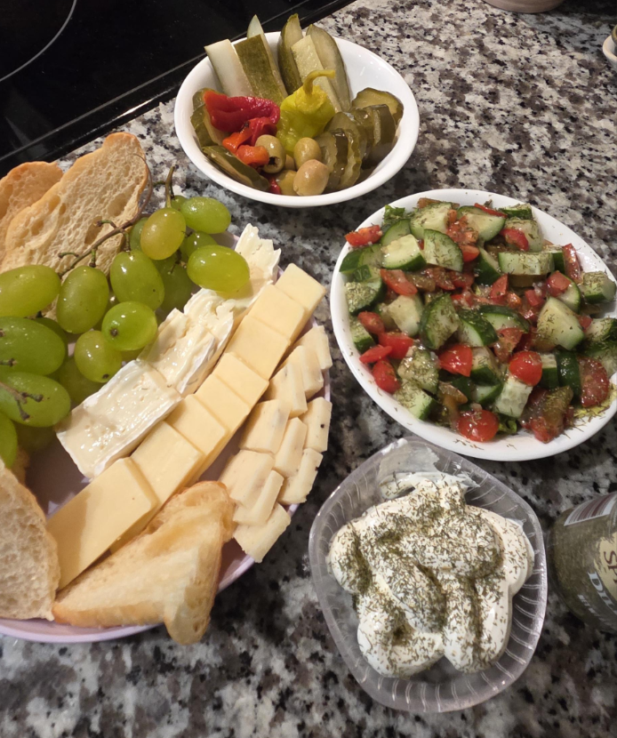 A spread of food on a granite countertop showcases how to stretch food—featuring sliced cheese, brie, green grapes, bread, pickles, olives, cucumber-tomato salad, and yogurt topped with dried herbs.