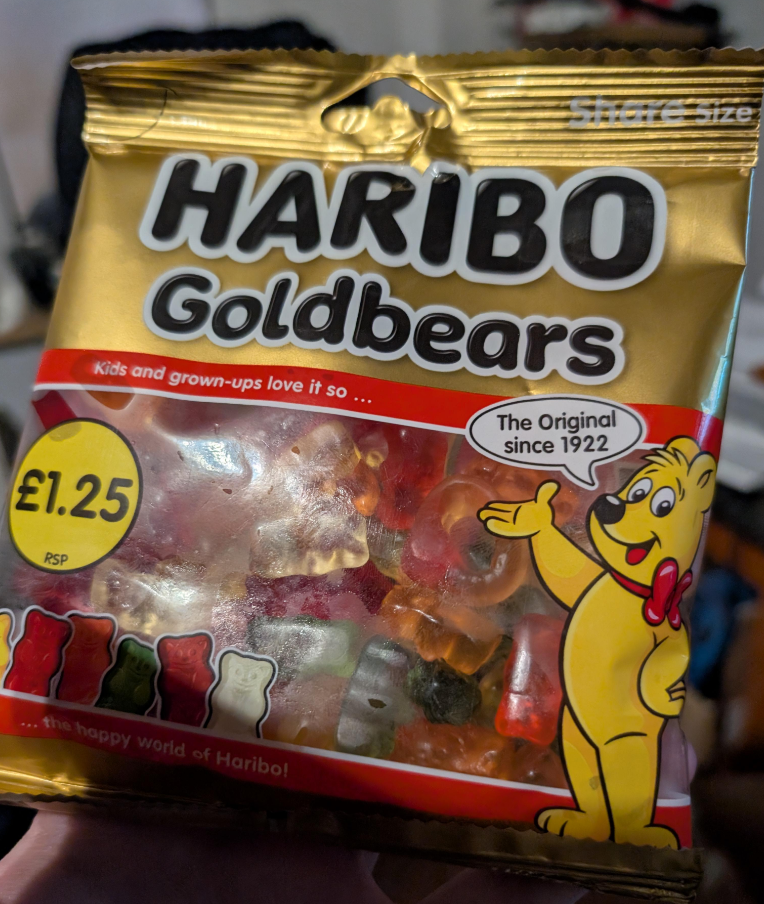 A hand holds a share-size bag of HARIBO Goldbears, widely known as the best gummy candy, showing colorful bears inside. The packaging features a yellow cartoon bear mascot and a price label of £1.25.