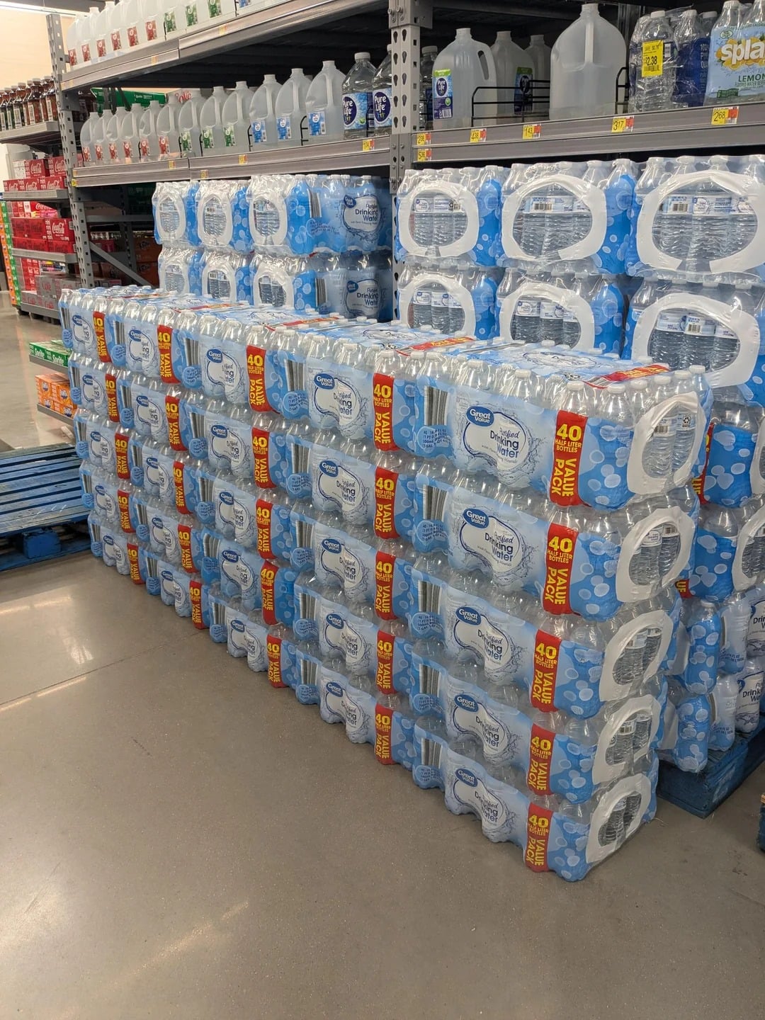 Cases of bottled water are stacked on blue pallets in a grocery store aisle, with shelves of gallon water jugs above and to the side. The clean, well-lit area highlights these popular Walmart products.