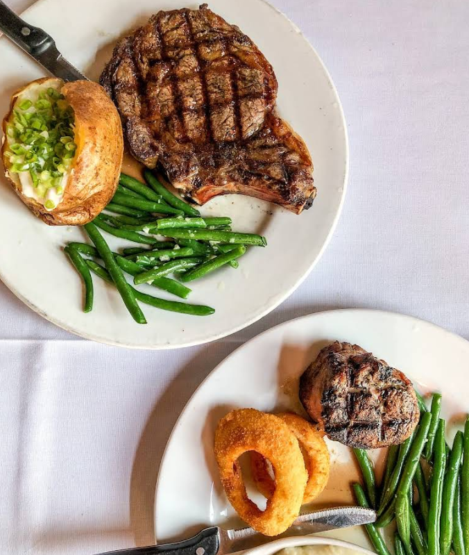 Two plates of food at a steakhouse on the west coast: one with grilled steak, green beans, and a baked potato topped with sour cream and chives; the other with filet mignon, green beans, and two crispy onion rings.