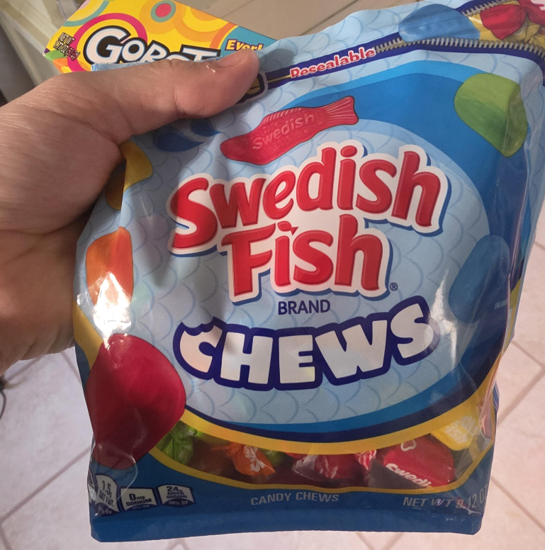 A hand holds a colorful bag of Swedish Fish Chews, the best gummy candy, featuring the red fish-shaped logo and bold red and white text on blue packaging. Part of a Go-Gurt box is visible in the background.