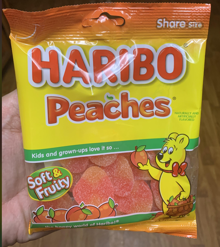 A hand holds a package of Haribo Peaches gummy candy. The yellow and orange bag, labeled "Soft & Fruity," features a cartoon bear with a peach and displays some of the best gummy candy shaped like peaches inside.