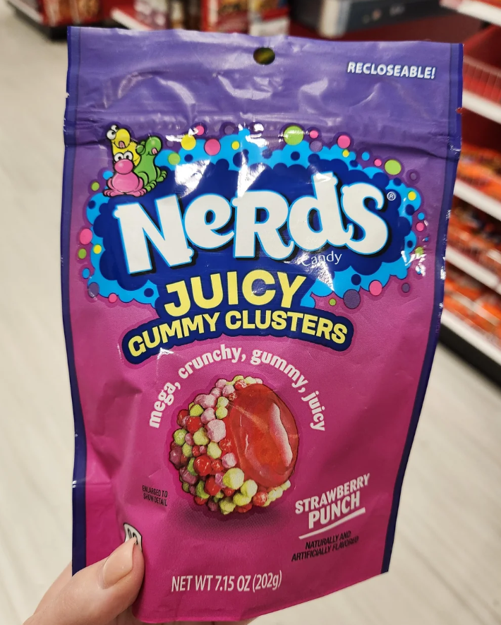 A hand holds a purple, resealable bag of Nerds Juicy Gummy Clusters in a store. The packaging advertises "Strawberry Punch" flavor and features a picture of the best gummy candy: colorful, crunchy gummy clusters.