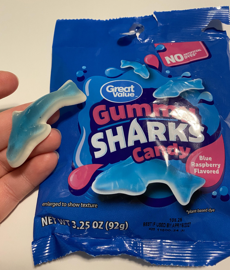 A hand holds a blue and white gummy candy shaped like a shark in front of a blue bag labeled "Great Value Gummy Sharks Candy, Blue Raspberry Flavored"—a contender for best gummy candy with no artificial dyes.