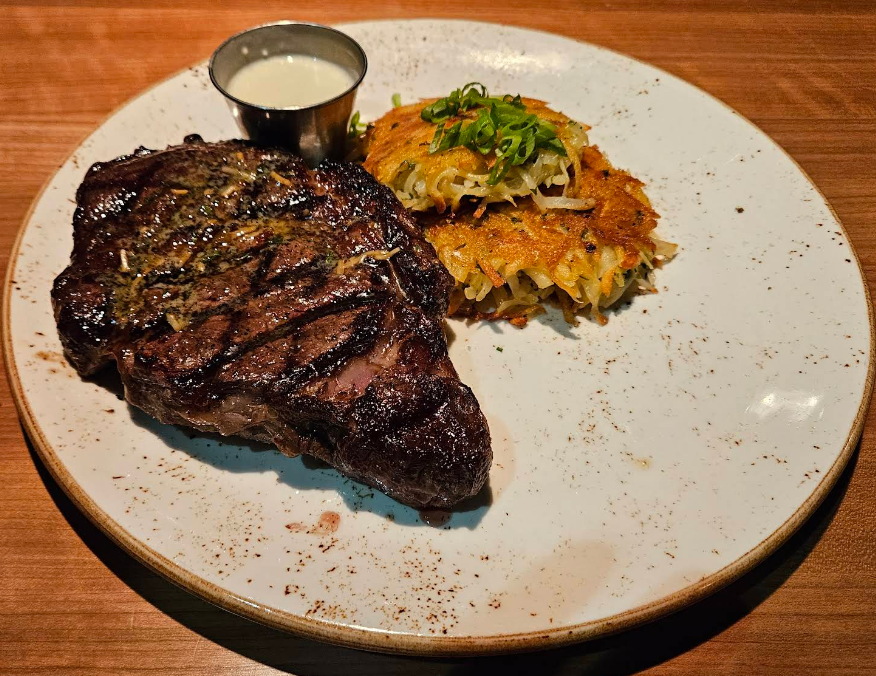 A grilled steakhouse on the West Coast steak with grill marks sits on a plate next to two hash brown patties topped with chopped green onions, plus a small cup of creamy white sauce, all served on a wooden table.