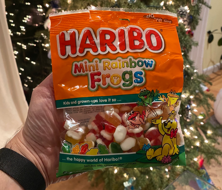 A hand holds a bag of Haribo Mini Rainbow Frogs, one of the best gummy candy treats, in front of a decorated Christmas tree. The orange package features colorful text and playful cartoon frog illustrations.