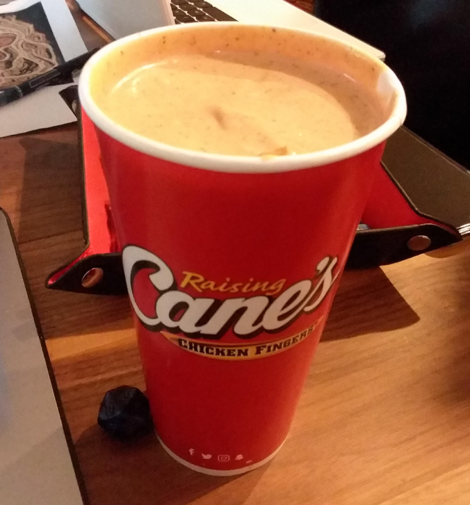 A large red Raising Cane's cup filled to the top with creamy fast-food sauces sits on a wooden table next to a laptop and a black item.