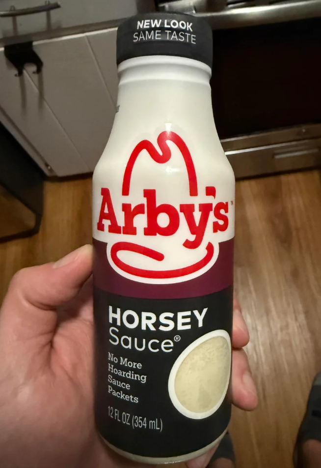 A hand holds a 12 fl oz bottle of Arby’s Horsey Sauce, one of the signature fast-food sauces, with the label stating “No More Hoarding Sauce Packets.” The bottle has a black cap and a red Arby’s logo. A kitchen appears in the background.