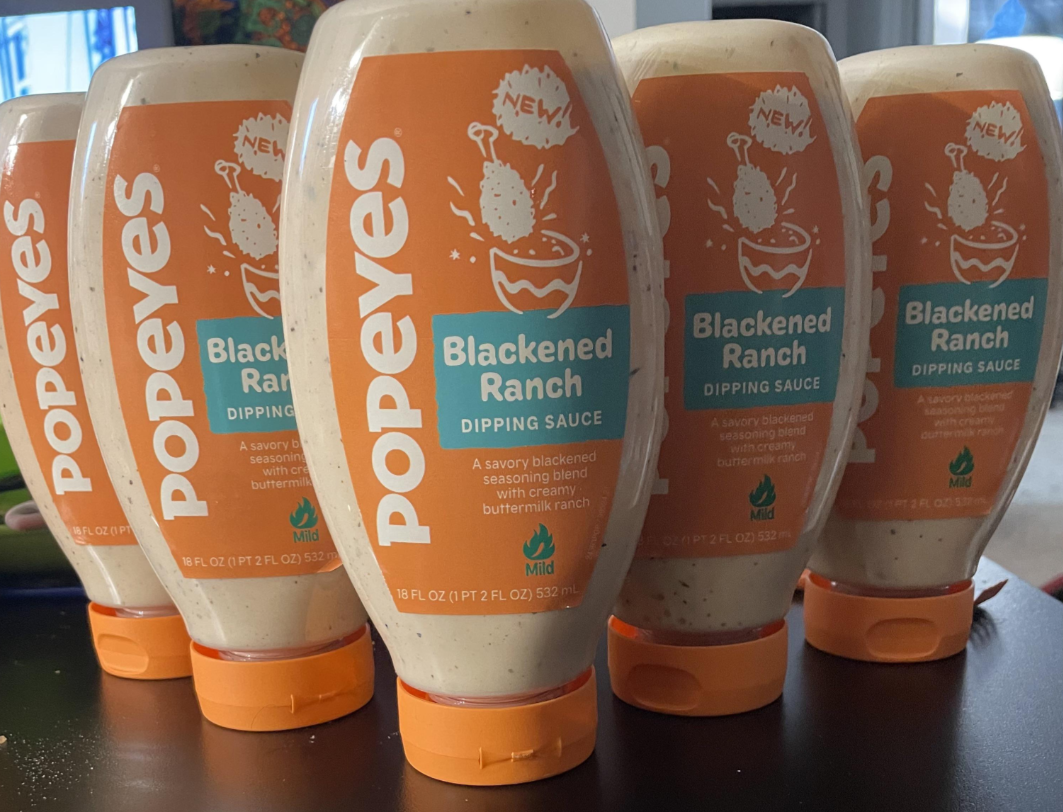 Five bottles of Popeyes Blackened Ranch, a favorite among fast-food sauces, are lined up on a dark surface, displaying their orange and white labels featuring a drumstick and sauce bowl.
