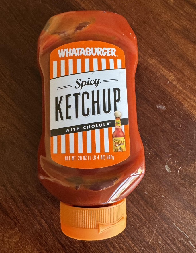 A 20-ounce bottle of Whataburger Spicy Ketchup with Cholula, a favorite among fast-food sauces, lies on a wooden surface. The bottle features an orange cap and a label with bold white and orange stripes.