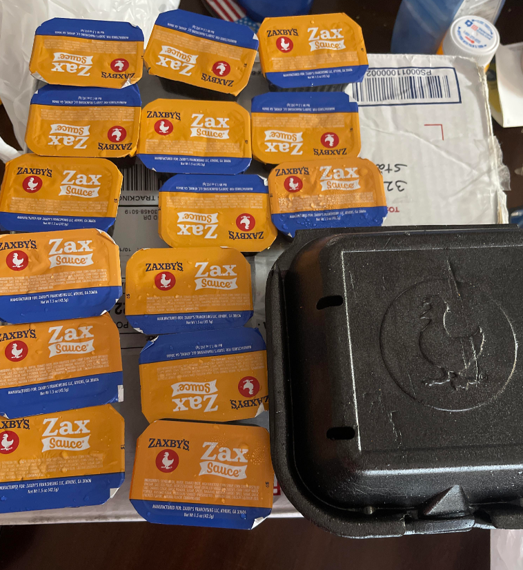 A group of Zaxby's fast-food sauces in Zax Sauce containers are arranged next to a closed black takeout container on top of a white mailing box.