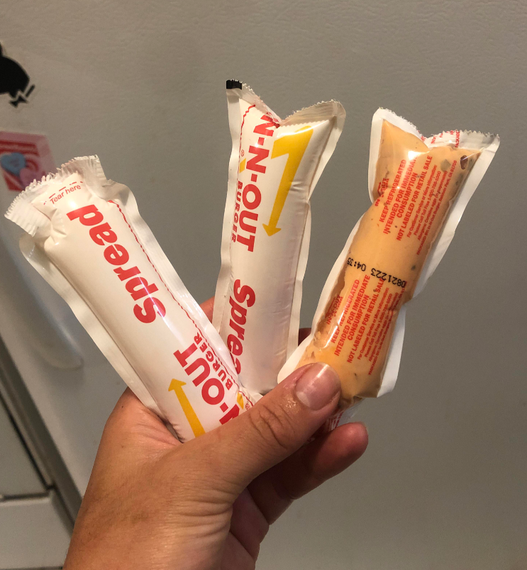 A hand holds three packets of In-N-Out Burger spread—iconic fast-food sauces—displayed in front of a white refrigerator. The white packets feature bold red and yellow text.
